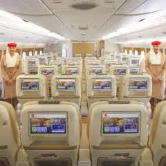 Uśmiechnięta stewardessa w klasie ekonomicznej Emirates. Rzędy wygodnych foteli z ekranami pokładowymi.