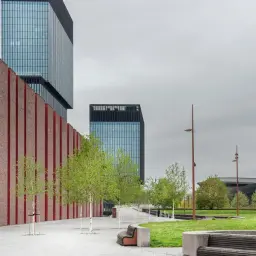 Nowoczesna architektura i zieleń tworzą ciekawe miejsca do 100 km od Katowic. Park z ławkami i drzewami przed szklanymi wieżowcami.