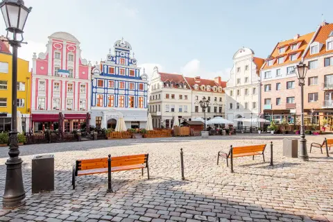 Kolorowy rynek miejski z kamienną nawierzchnią, ławkami i zabytkowymi kamienicami.