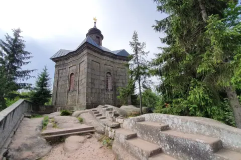 Kamienna kaplica na skale, otoczona drzewami. Broumowskie ściany budowli wznoszą się ku niebu, a kamienne schody prowadzą do jej wnętrza.