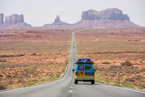 Błękitny van z bagażem na dachu jedzie pustynną drogą w USA. Monument Valley w tle. To część ich wycieczki do USA.