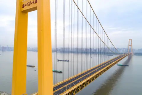 Żółty, wiszący most w Chinach przecina szeroką rzekę, po której płyną barki. W oddali widać panoramę miasta.