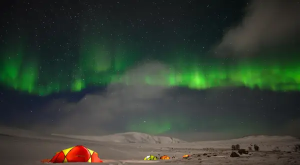 Nocne niebo nad Norwegią rozświetla zorza polarna. Kilka kolorowych namiotów rozbitych na śniegu tworzy przytulne schronienie pod tym niezwykłym zjawiskiem.