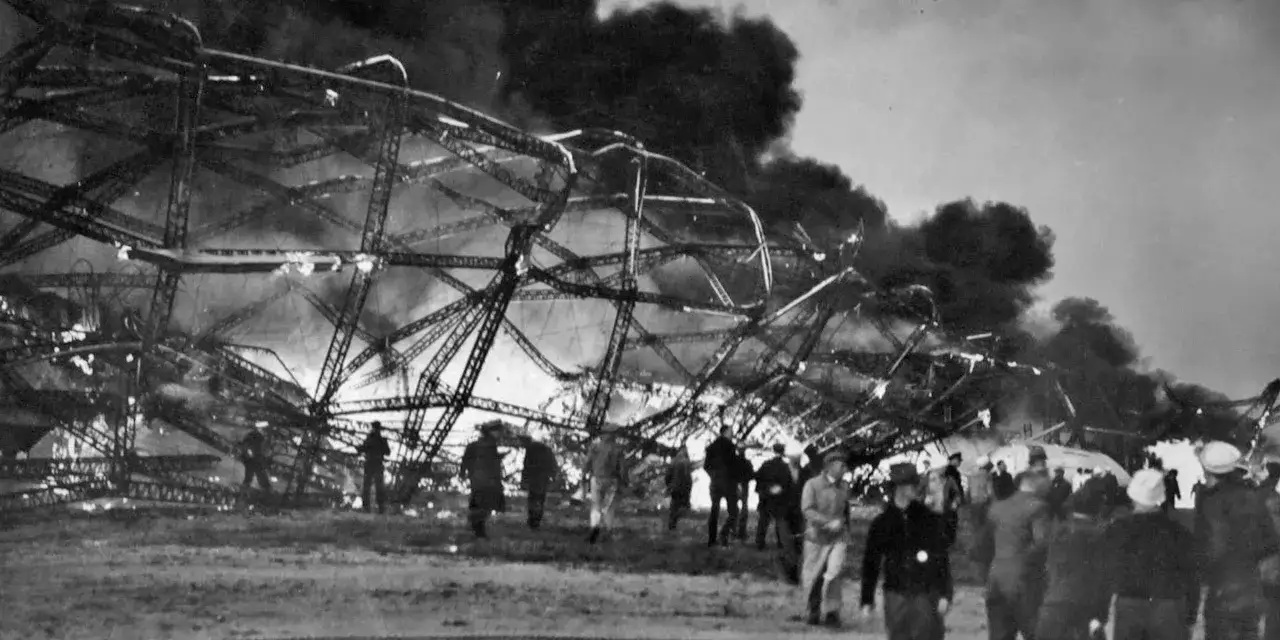 Zniszczony szkielet sterowca Hindenburg płonie, otoczony dymem. Ludzie obserwują tragedię z ziemi.