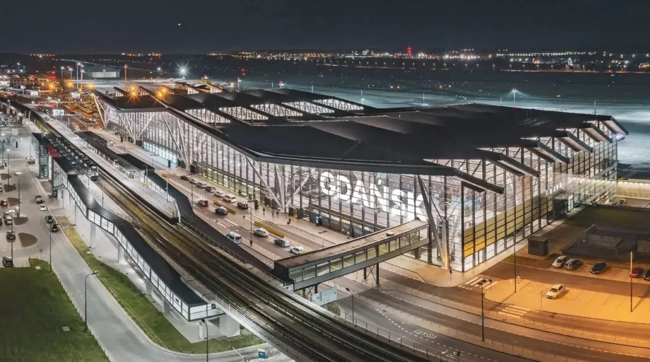 Nowoczesny terminal lotniska w Gdańsku nocą, największe lotnisko w Polsce, z widocznymi światłami miasta w tle.
