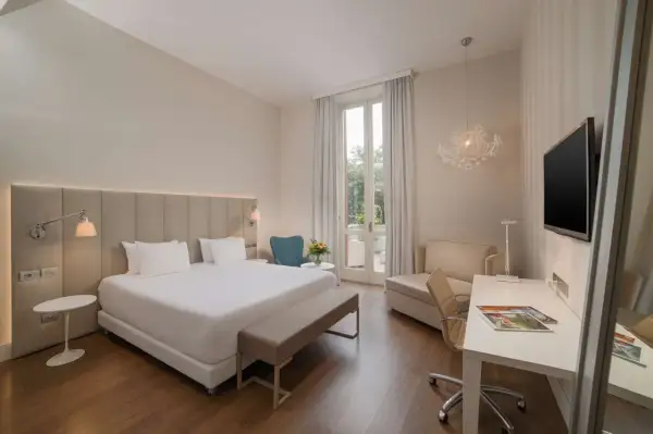 Elegancki pokój hotelowy z dużym łóżkiem, biurkiem i fotelem. Widok na taras z zielenią. Idealne miejsce na wypoczynek w kurorcie na riwierze francuskiej.