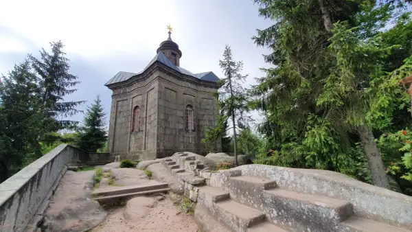 Kamienna kaplica na skale, otoczona drzewami. Broumowskie ściany budowli wznoszą się ku niebu, a kamienne schody prowadzą do jej wnętrza.