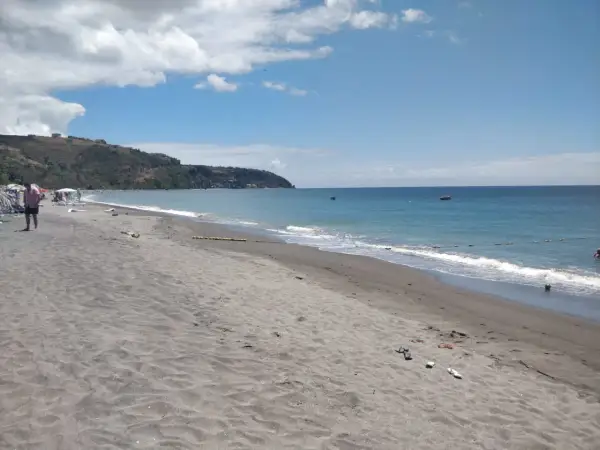 Plaża na wyspie Dominika z ciemnym piaskiem, turkusową wodą i zielonymi wzgórzami w tle.