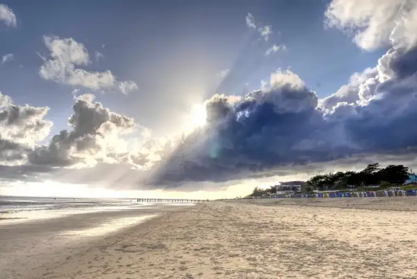 Plaża na wyspie w Niemczech. Słońce przebija się przez burzowe chmury, oświetlając piaszczysty brzeg i rzędy kolorowych budek plażowych.