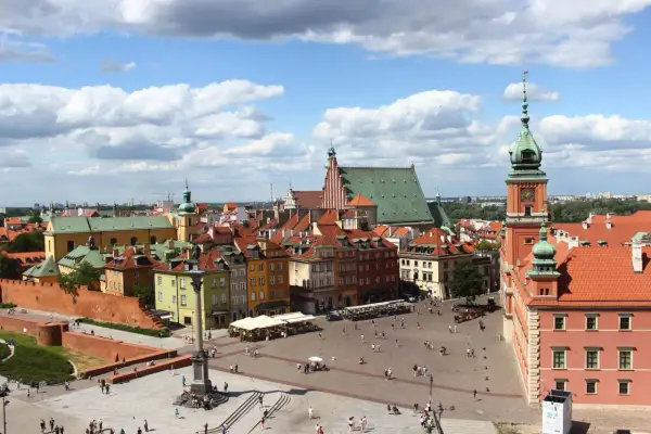 Widok na Zamek Królewski i Kolumnę Zygmunta III Wazy na placu w warszawskiej starówce. Tłumy spacerują po bruku, a słońce przebija się przez chmury.