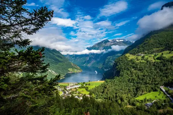 Widok na fiord podczas rejsu do Norwegii. Statek wycieczkowy płynie po spokojnej wodzie, otoczonej zielonymi górami i chmurami.