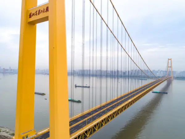 Żółty, wiszący most w Chinach przecina szeroką rzekę, po której płyną barki. W oddali widać panoramę miasta.